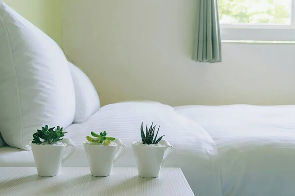 A neatly made bed with white bedding, three small potted plants on a white table, and a window with a curtain.