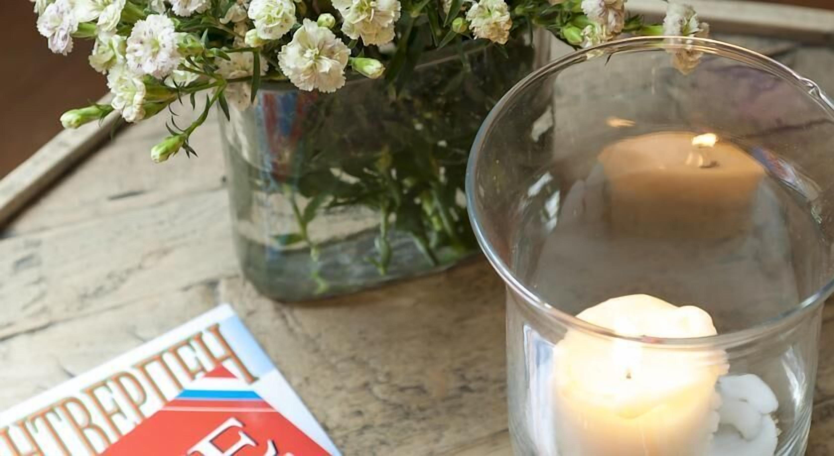 Une bougie dans un bocal en verre, un vase avec des fleurs et un livre sur une surface en bois.