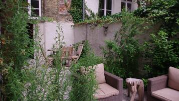 Un espace de détente extérieur accueillant, avec des meubles en osier et un tabouret en bois rustique, entouré d’une végétation luxuriante et d’un mur de briques.