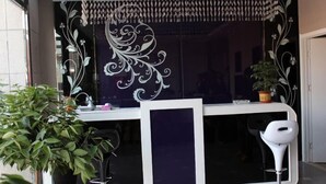 Un mostrador de recepción de diseño moderno, con un panel azul oscuro y detalles blancos. A la izquierda hay una planta en maceta y a la derecha una silla blanca.