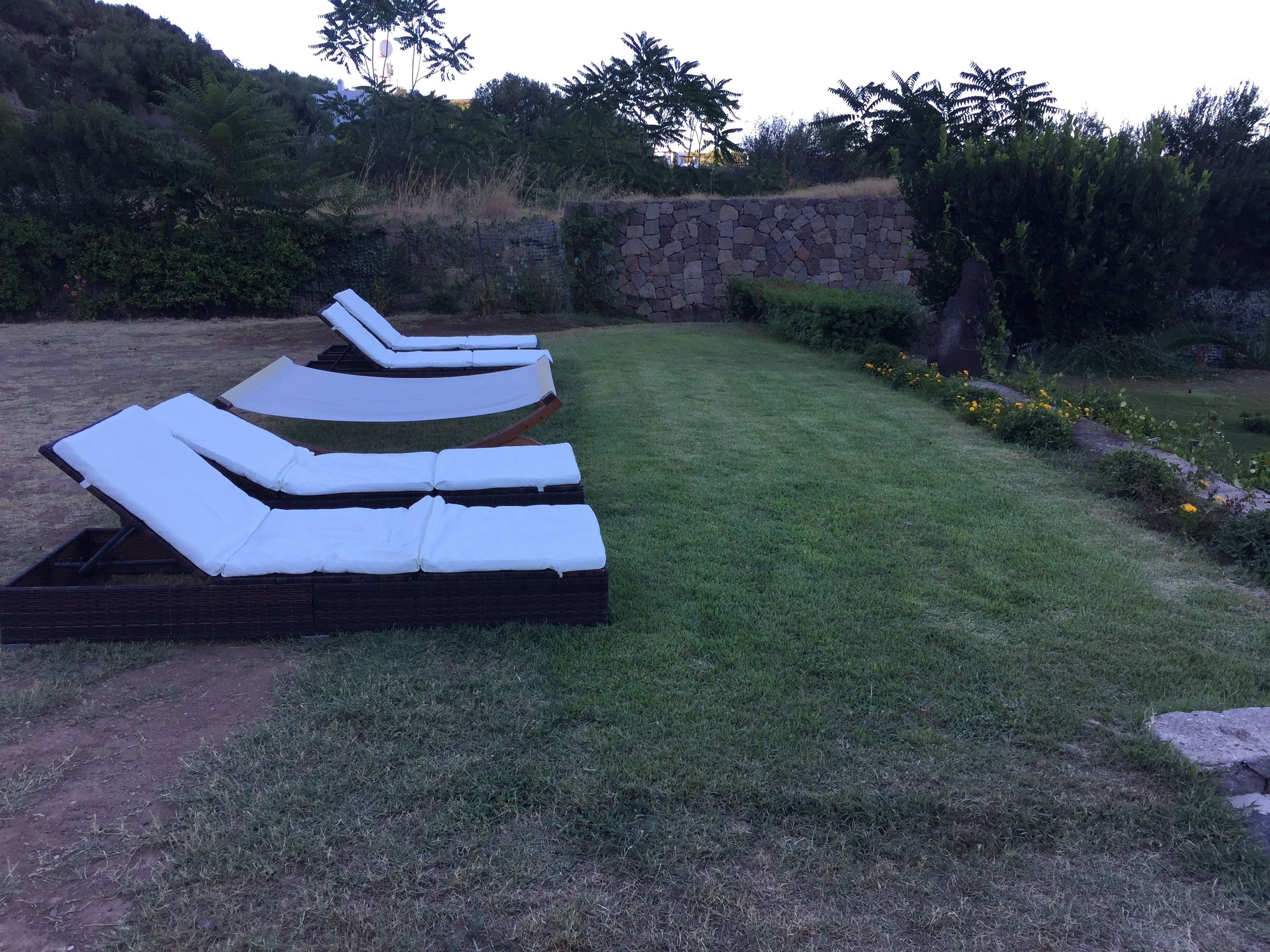 Una fila de sillas de exterior con cojines blancos, dispuestas sobre un césped con un muro de piedra y árboles al fondo.