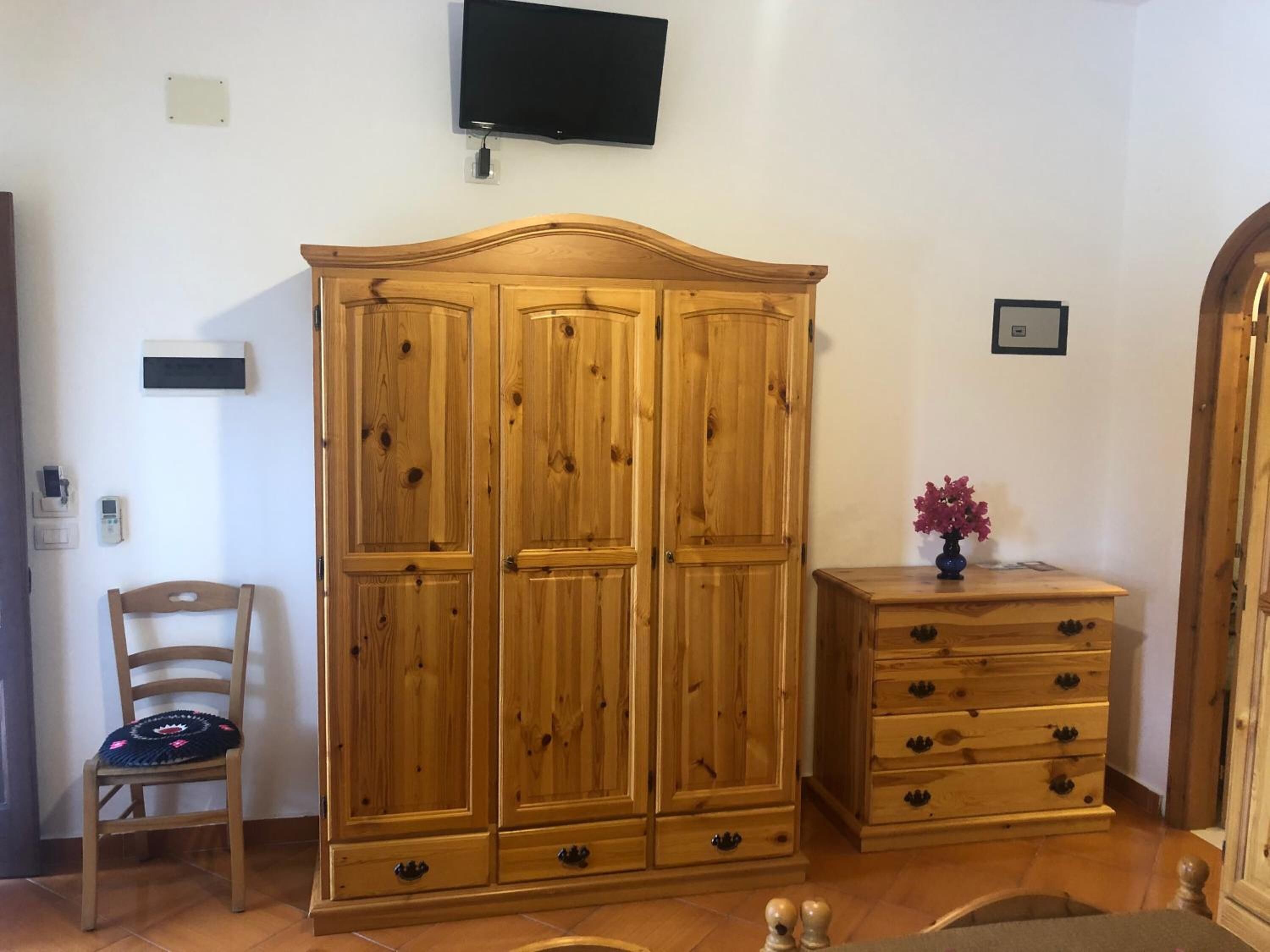 A room with a wooden wardrobe, a chair, a small table with a vase, and a television mounted on the wall.