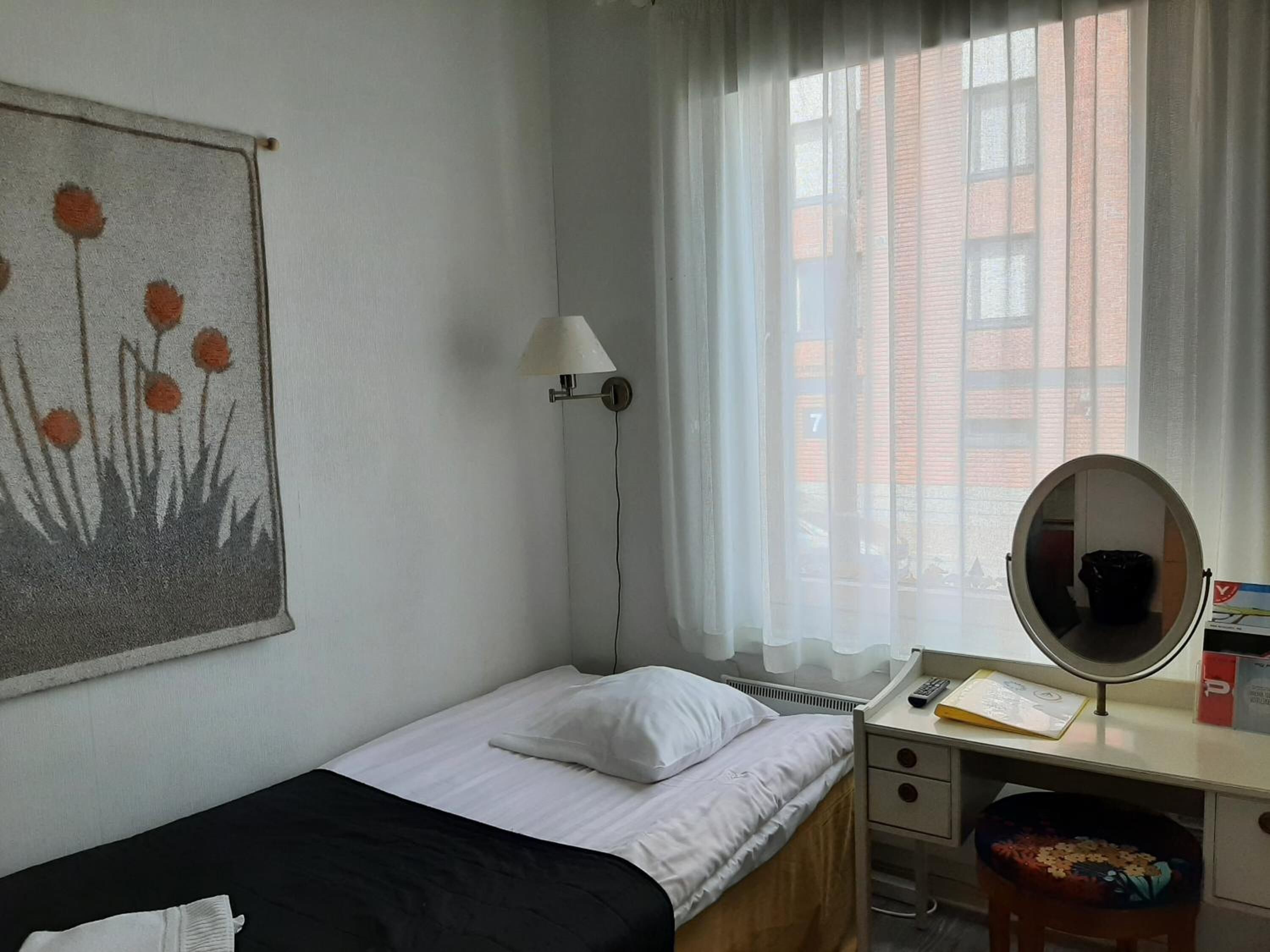 Quarto pequeno e bem arrumado, com uma cama de solteiro, uma escrivaninha com espelho e uma janela com cortinas translúcidas.