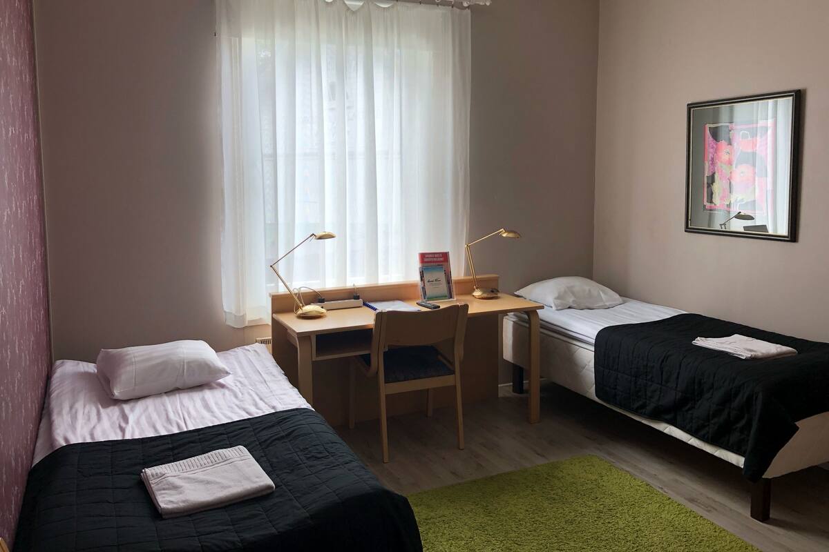 A hotel room with two single beds, a desk with two lamps, a chair, and a green rug.