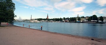 Näkymä joen varrelta, jossa on hiekkaranta, veneitä ja taustalla näkyy selvästi kirkon torni.