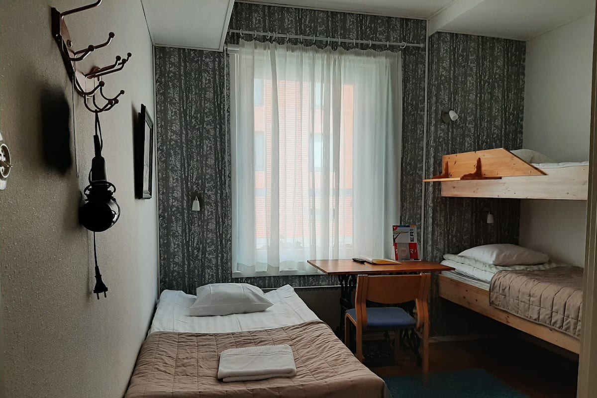 A compact room with a bunk bed, a single bed, a desk with a chair, a window with sheer curtains, and a wall-mounted lamp.