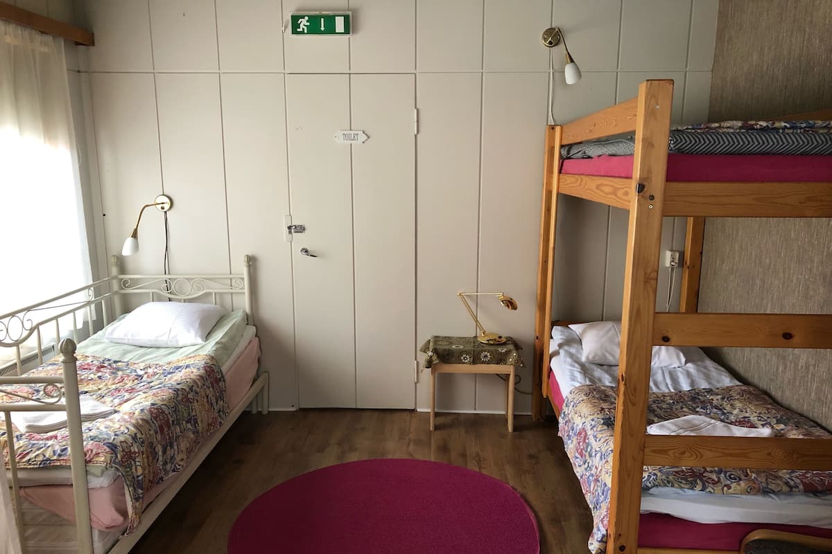 A dormitory room with two bunk beds, a small bedside table with a lamp, a red rug, and a door with a green exit sign.