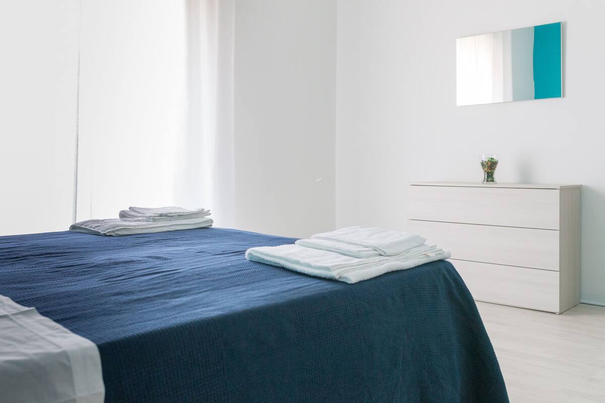 Un lit bien fait, avec une literie bleue, une commode blanche et un petit vase posé sur la commode.