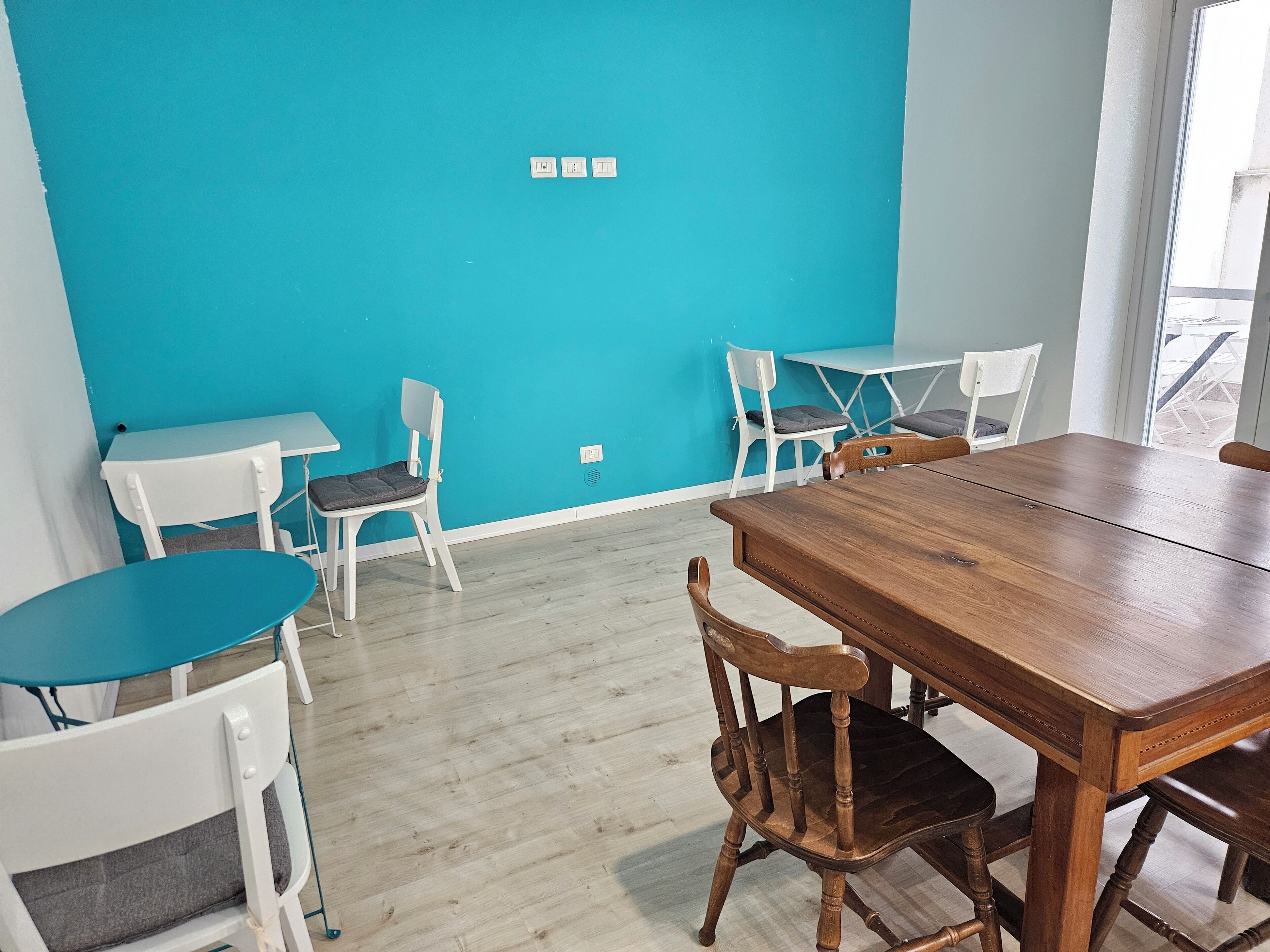 A room with a teal wall, wooden dining table, white chairs, and a window with a view.