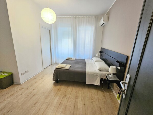A hotel room with a bed, bedside lamp, air conditioning unit, and a view of the outdoors through sheer curtains.