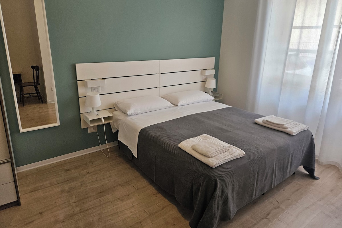 Une chambre d’hôtel bien aménagée, avec un grand lit, une table de chevet avec une lampe, un miroir et une fenêtre ornée de rideaux légers.