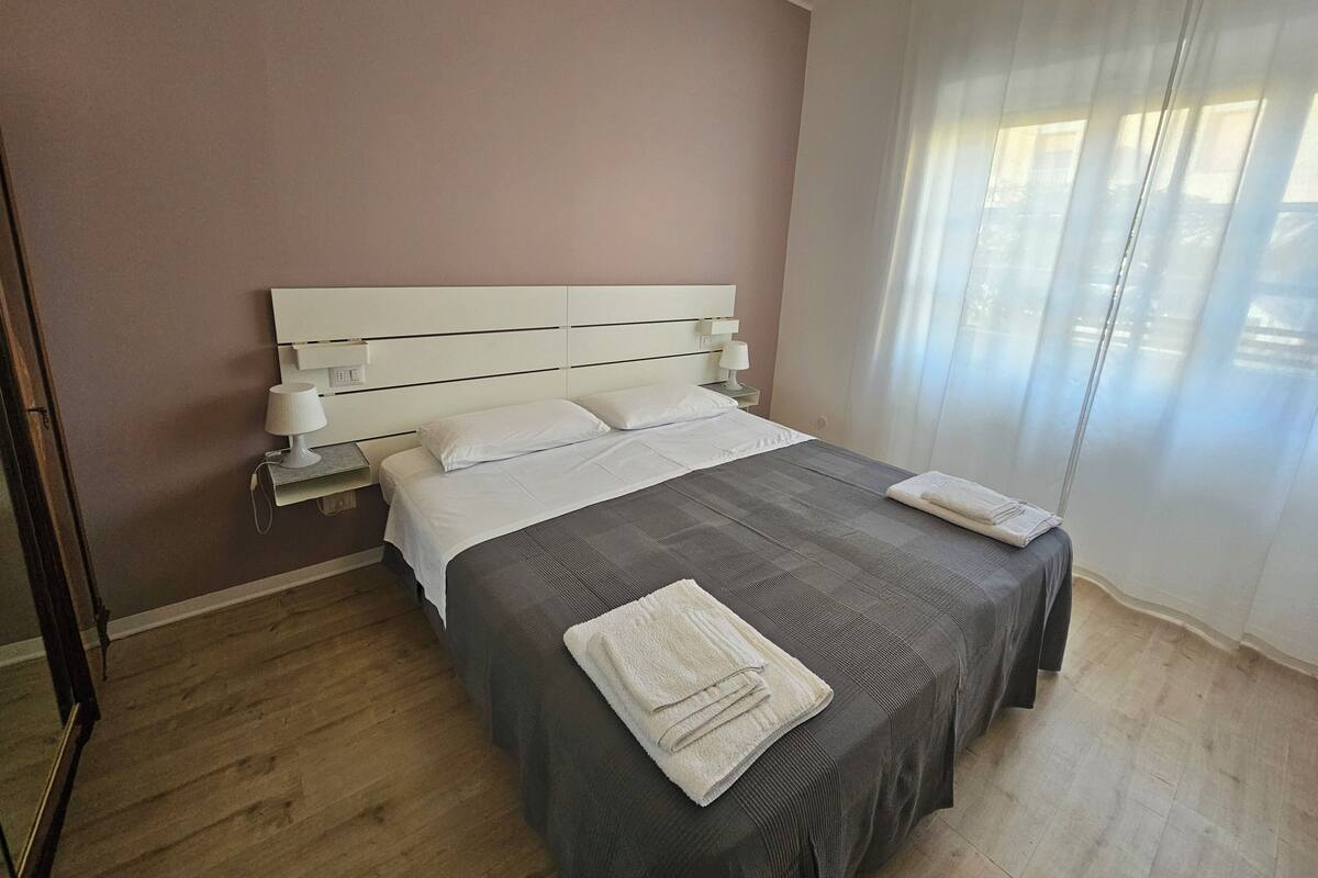 Un lit bien fait, avec du linge de lit blanc et une couvre-lit grise, une table de chevet avec une lampe, et une fenêtre ornée de rideaux légers.