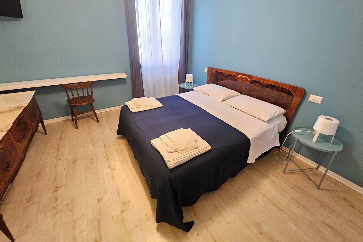 Un lit bien fait, recouvert d’une couvre-lit bleu foncé et agrémenté d’oreillers blancs, une chaise en bois, une petite table avec une lampe et une fenêtre avec des rideaux.