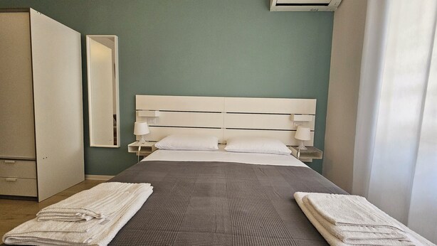 Un lit bien fait, recouvert d’une couverture grise, avec deux oreillers blancs et deux serviettes pliées de chaque côté. On y trouve une table de chevet blanche avec une lampe, un climatiseur fixé au mur et un miroir sur le mur.