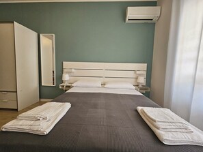 A neatly made bed with a grey bedspread, two white pillows, and two folded towels on either side. There is a white bedside table with a lamp, a wall-mounted air conditioning unit, and a mirror on the wall.