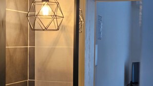 Une salle de bain aux murs recouverts de carreaux foncés, dotée d’un luminaire suspendu aux formes géométriques et d’un miroir à cadre noir.