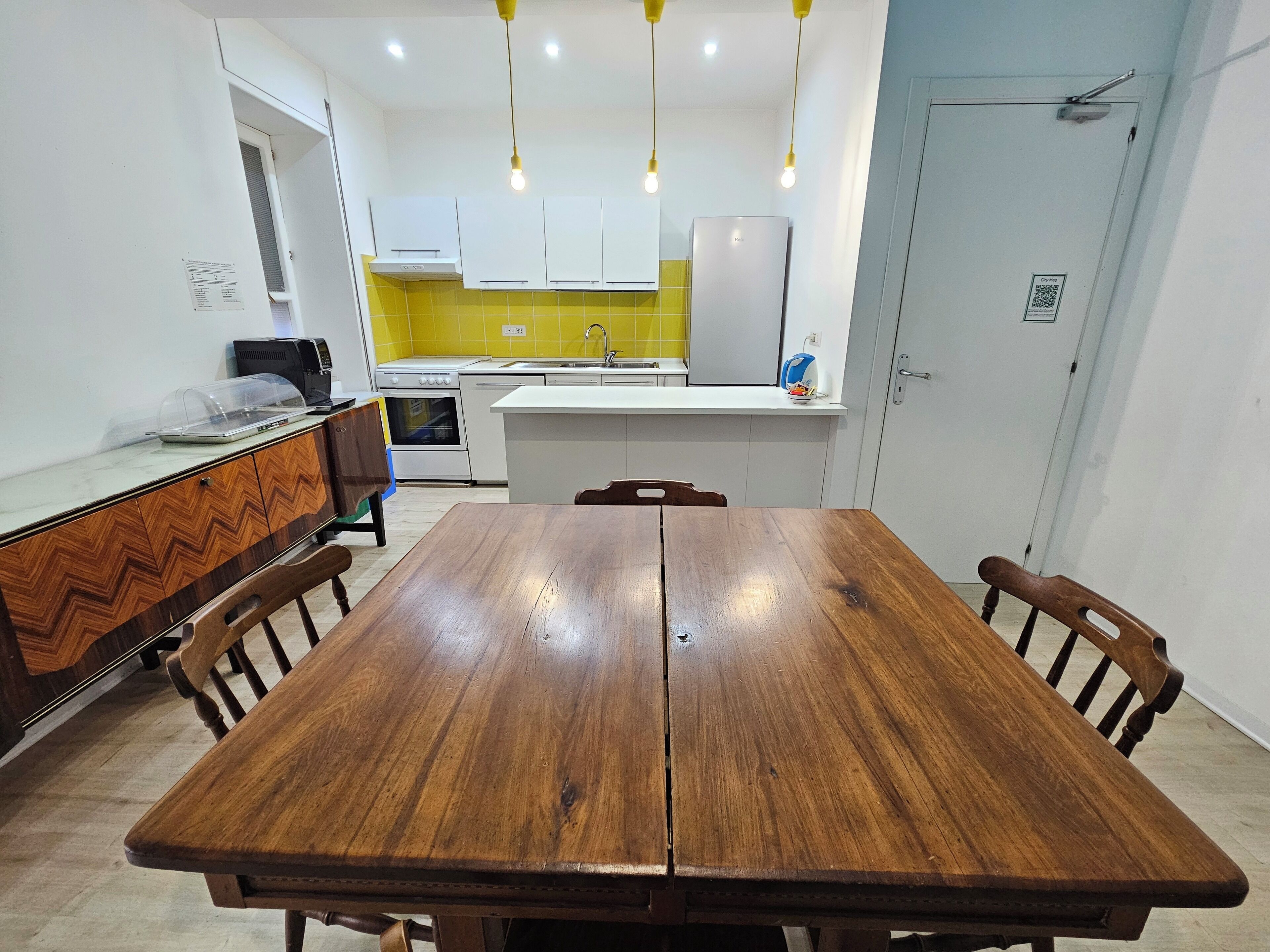 A dining table with chairs, a kitchen area with white cabinets and a yellow backsplash, and a door with a QR code.