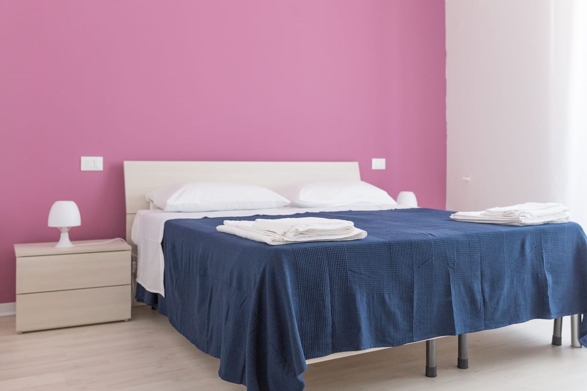 Un lit bien fait, recouvert d’une couvre-lit bleue, une table de chevet blanche avec une lampe, et un mur rose.