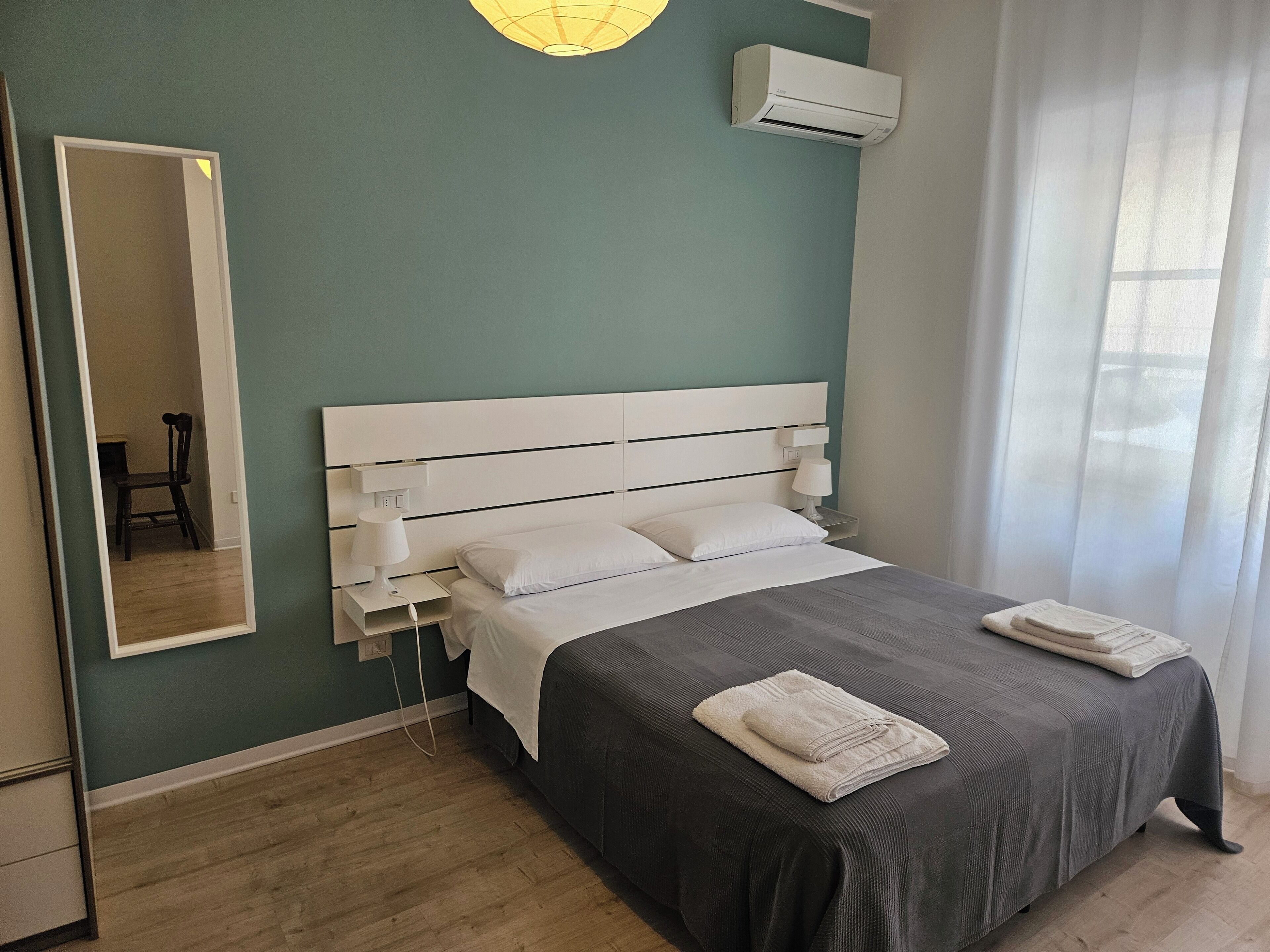 A bedroom with a bed, bedside tables, a mirror, an air conditioning unit, and a window with curtains.