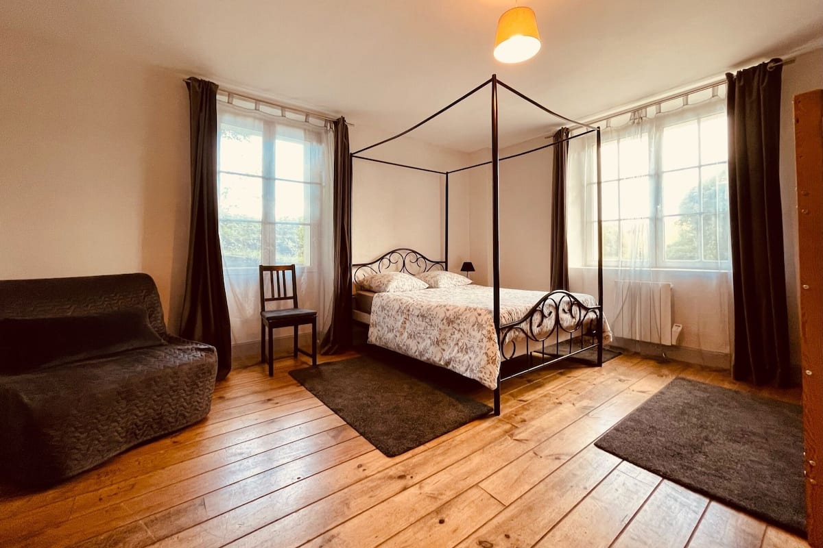 A four-poster bed with a floral bedspread, a wooden floor, a chair, and a sofa.