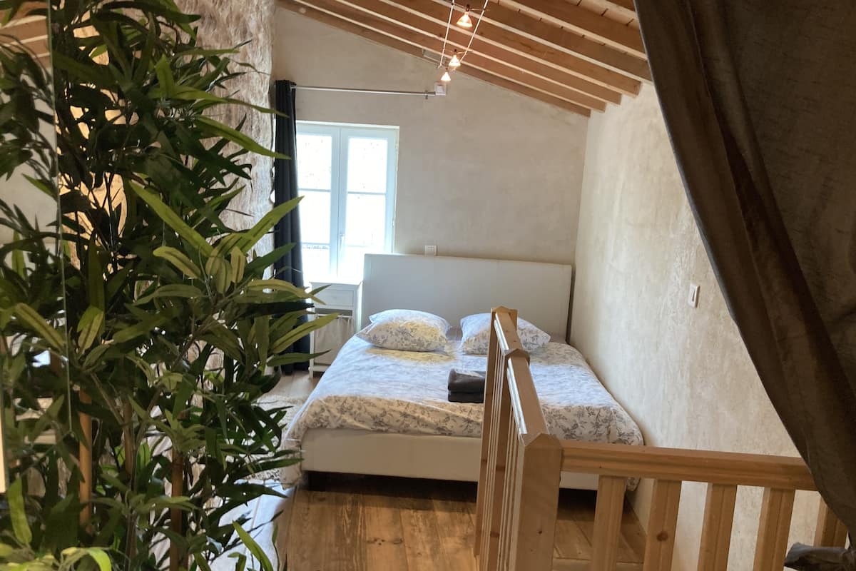 A bedroom with a bed, a nightstand, a window, and a plant.