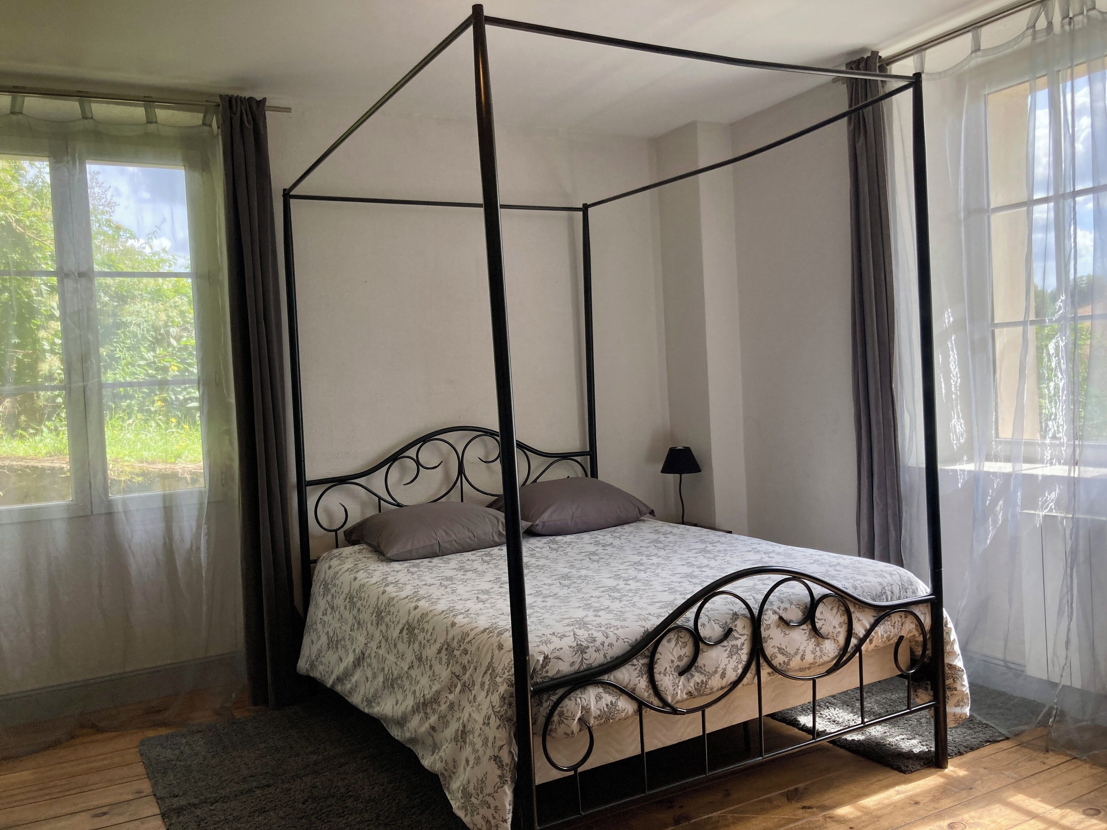 Ein Himmelbett mit floralem Bettbezug und zwei Kissen, platziert in einem Raum mit Holzboden und großen Fenstern mit durchsichtigen Vorhängen.