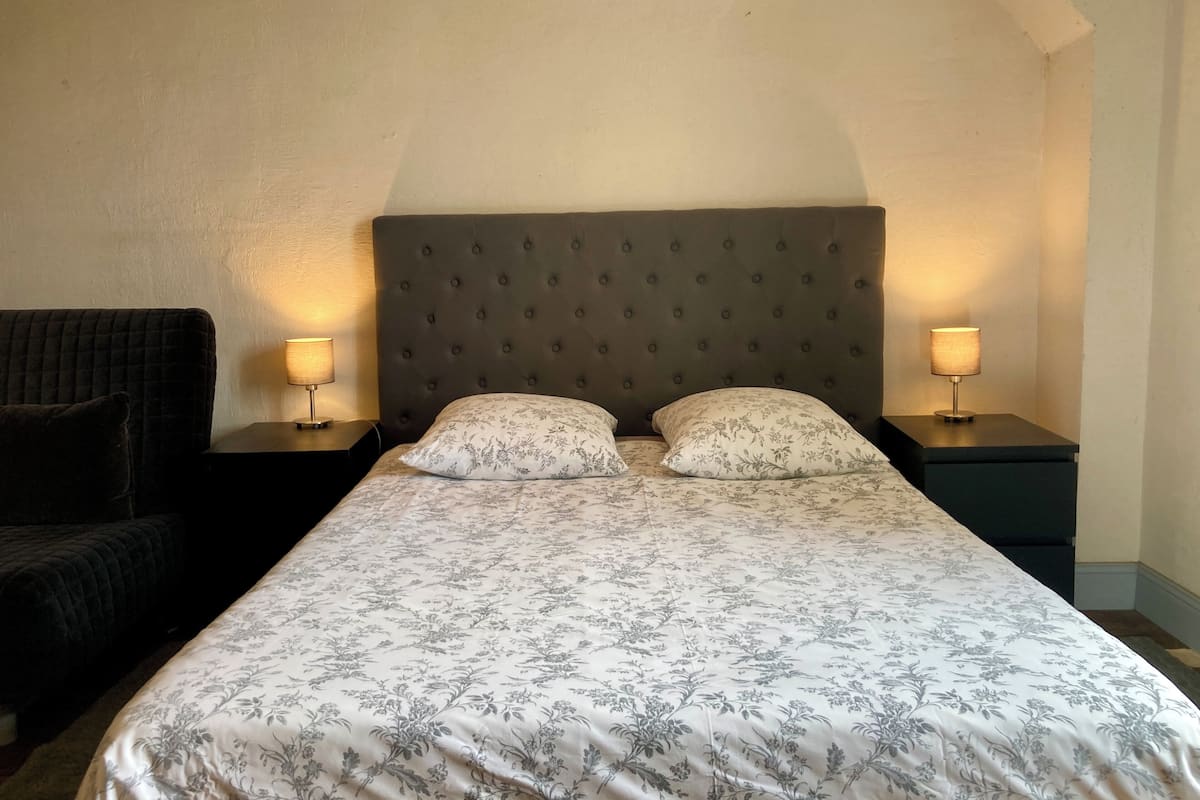 A bedroom with a large bed, two bedside lamps, a dark armchair, and a plain wall.