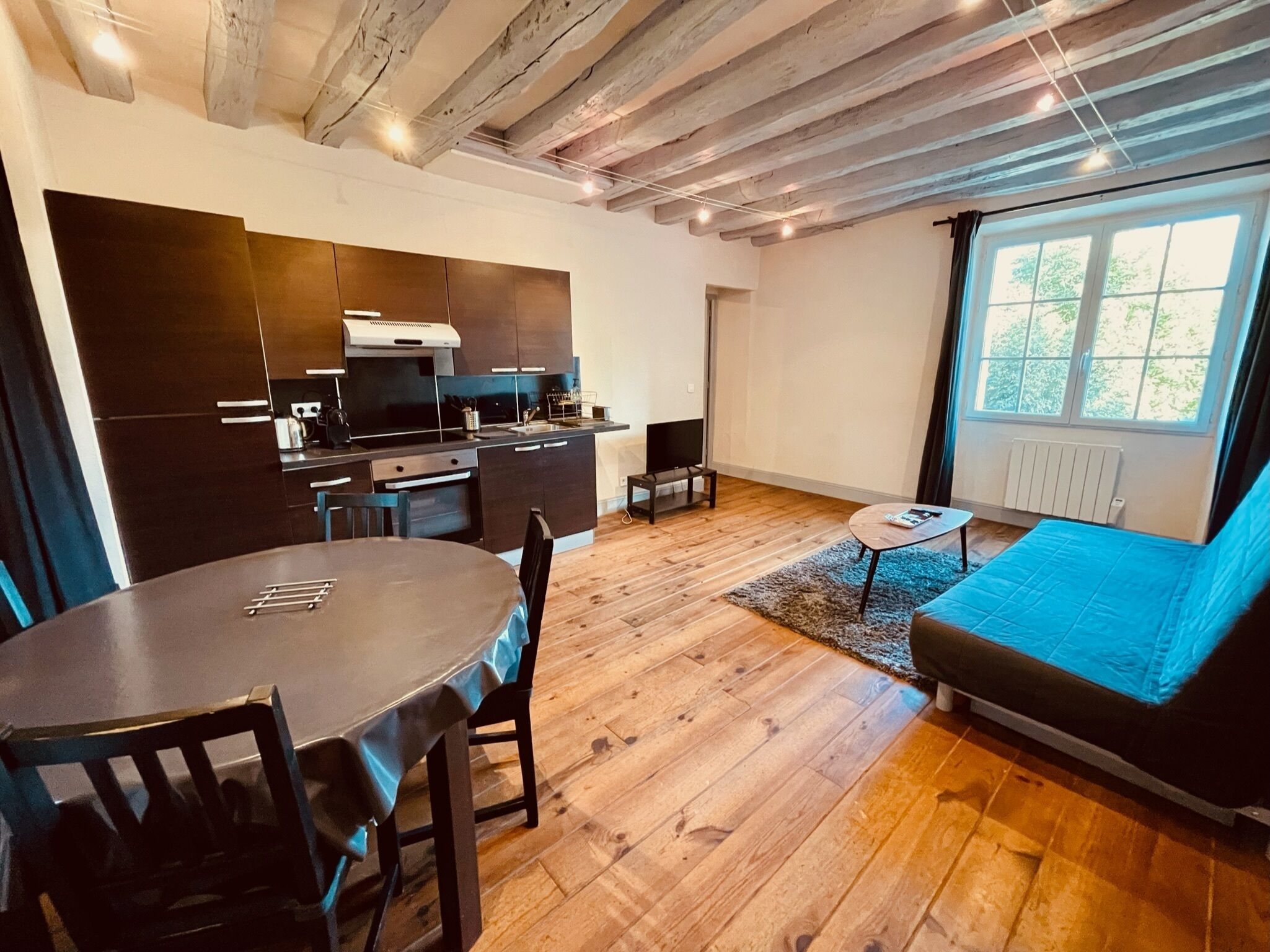 A modern kitchen with dark cabinets and a wooden ceiling, a dining area with a round table and chairs, and a blue sofa in a room with wooden flooring and a large window.