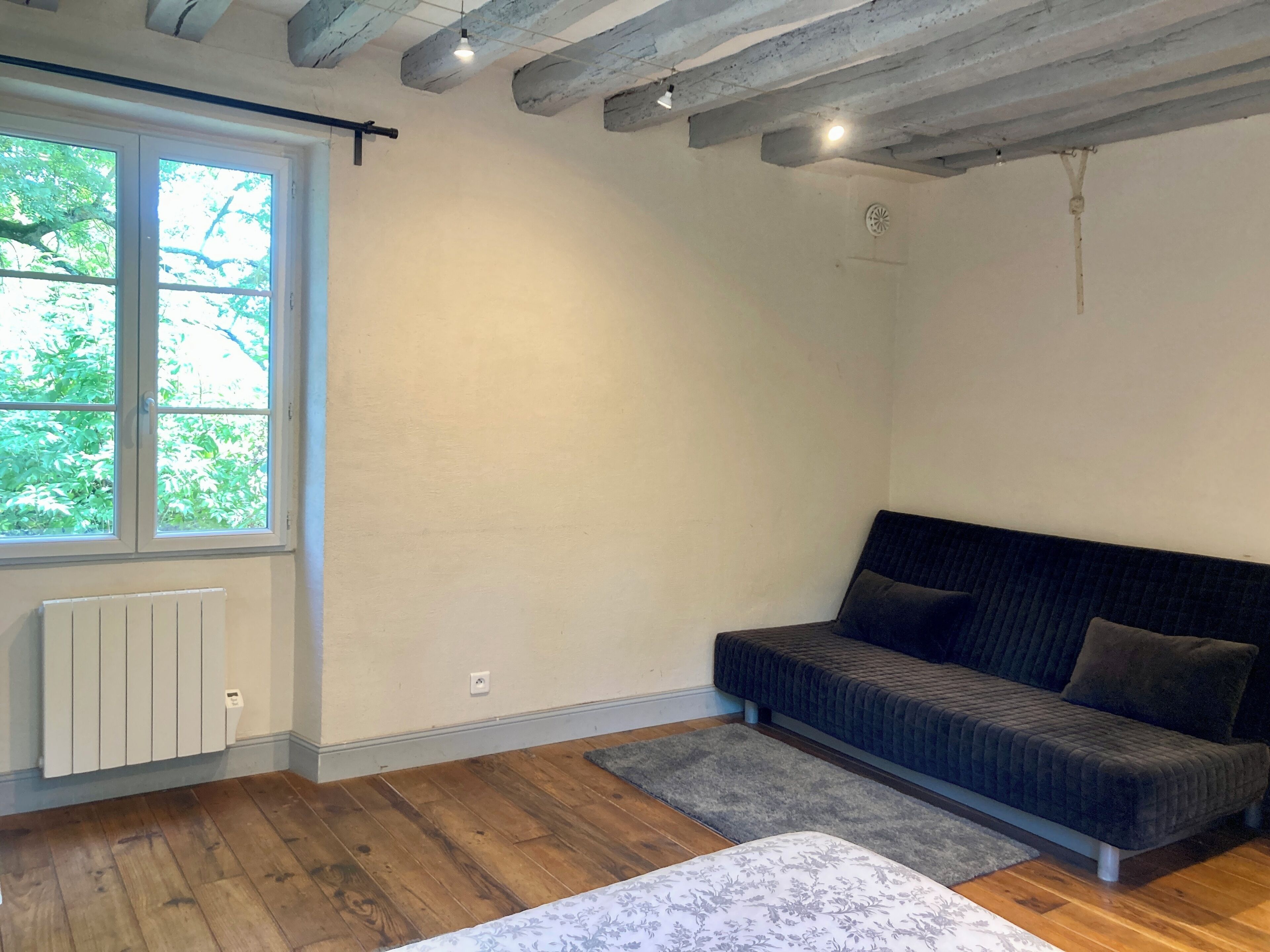 Een kamer met een groot raam, een radiator, een grijze bank en een houten vloer.