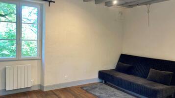 Een kamer met een groot raam, een radiator, een grijze bank en een houten vloer.