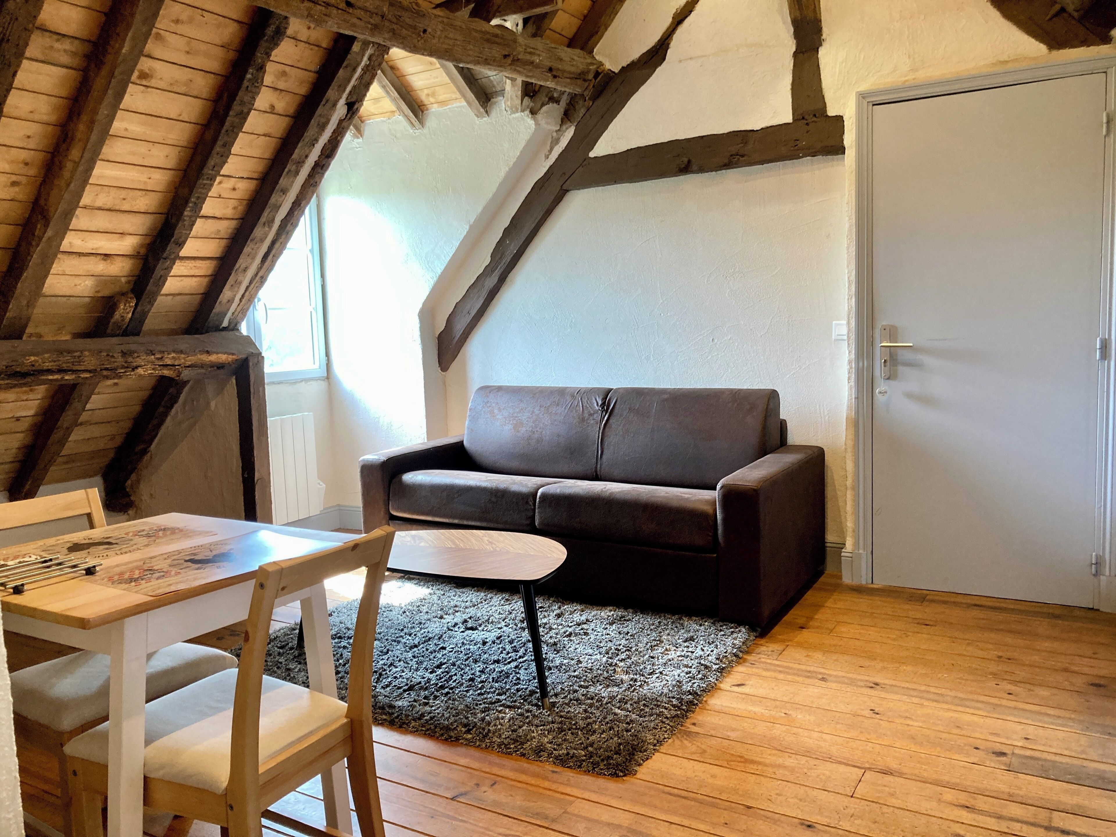 Een knus loft slaapkamer met een houten plafond, een donkerbruine bank, een tafeltje en een stoel.