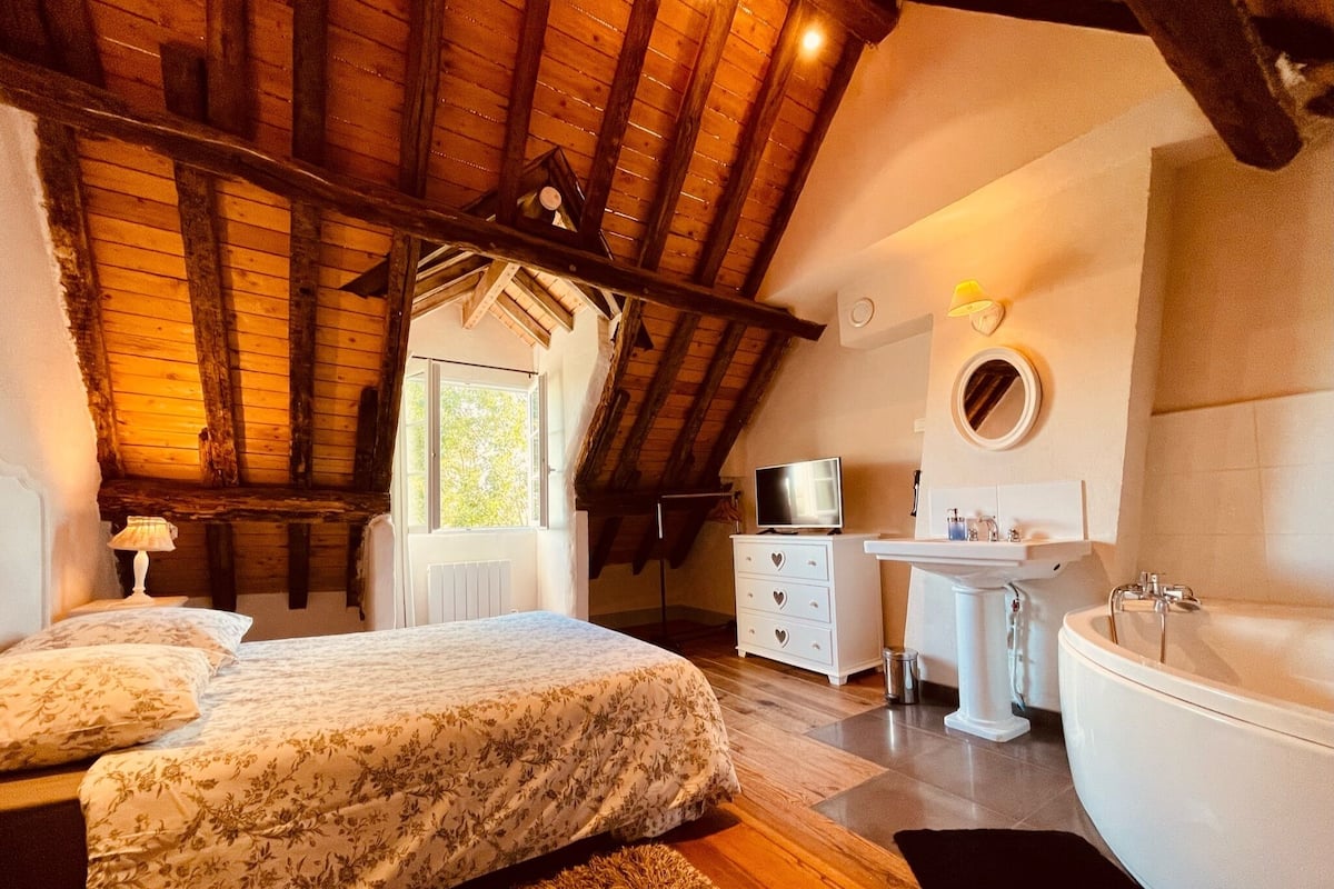 A bedroom with a large bed, a dresser, a TV, and a bathroom with a bathtub and sink.