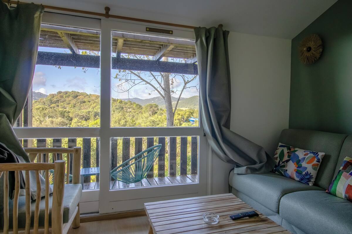 A room with a view of a forested area, a wooden chair, a sofa, and a table with a remote control and a glass.