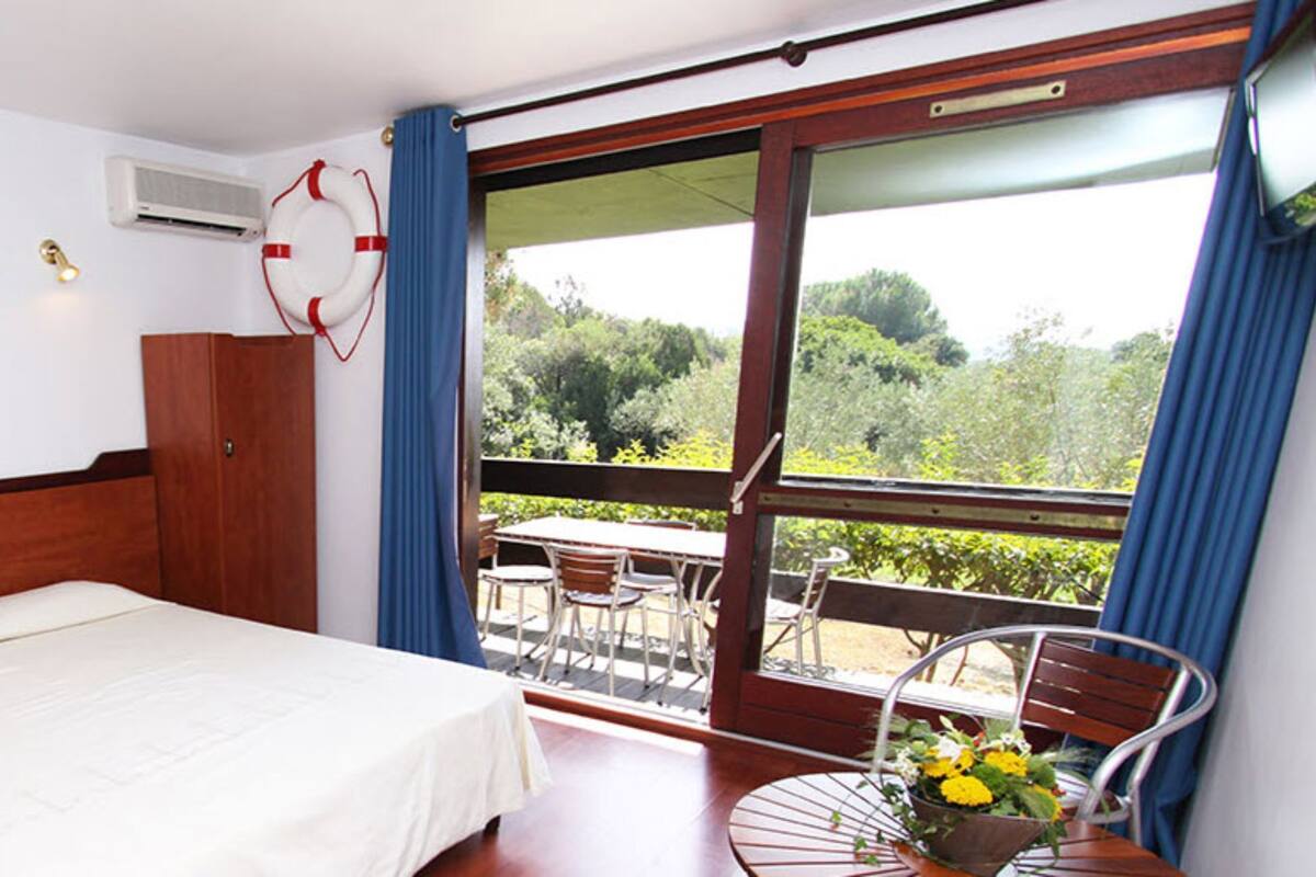 A hotel room with a bed, a desk, a chair, a table with flowers, a window with a view of greenery, and a life preserver on the wall.