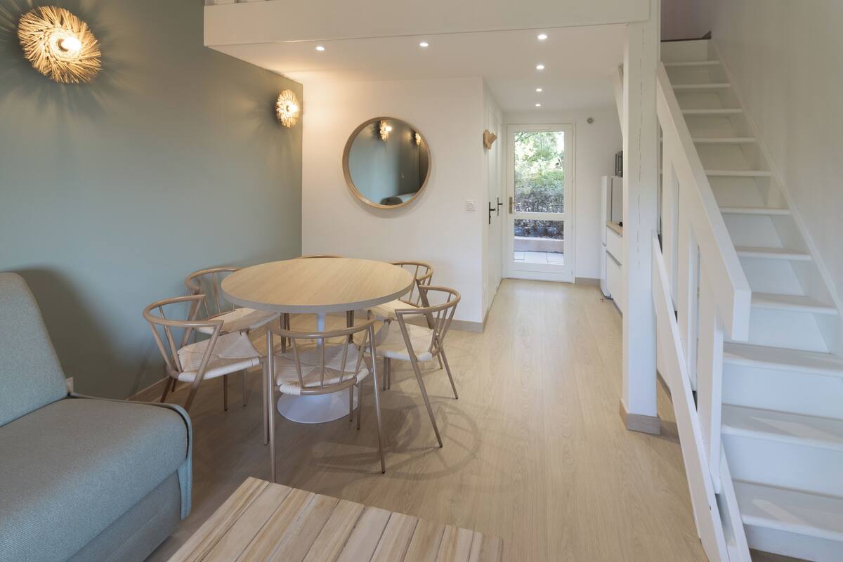 A modern living room with a round table, chairs, a sofa, a staircase, and a view of the outdoors.