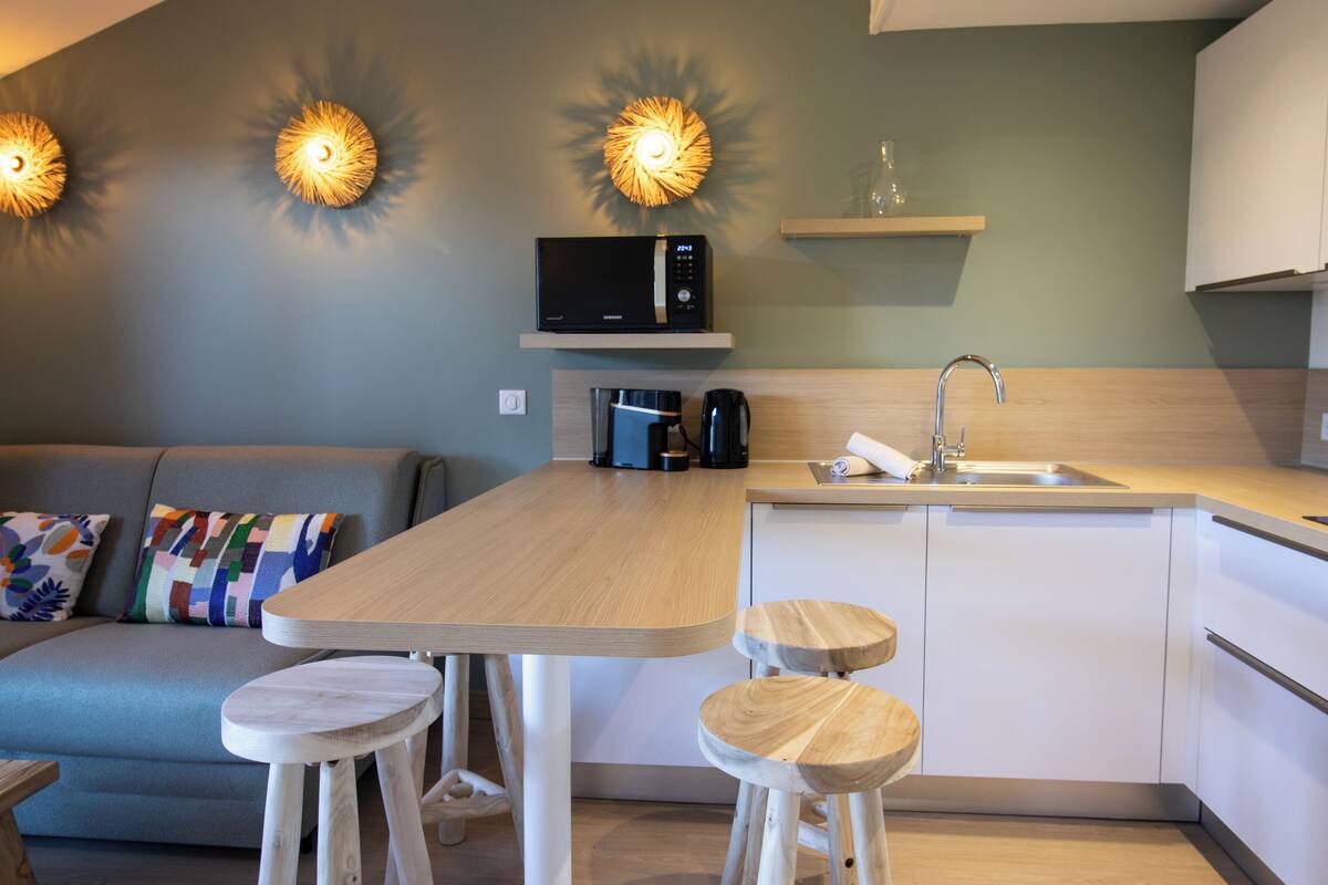 A compact kitchen with a microwave, coffee maker, and sink. There are two stools and a small table.