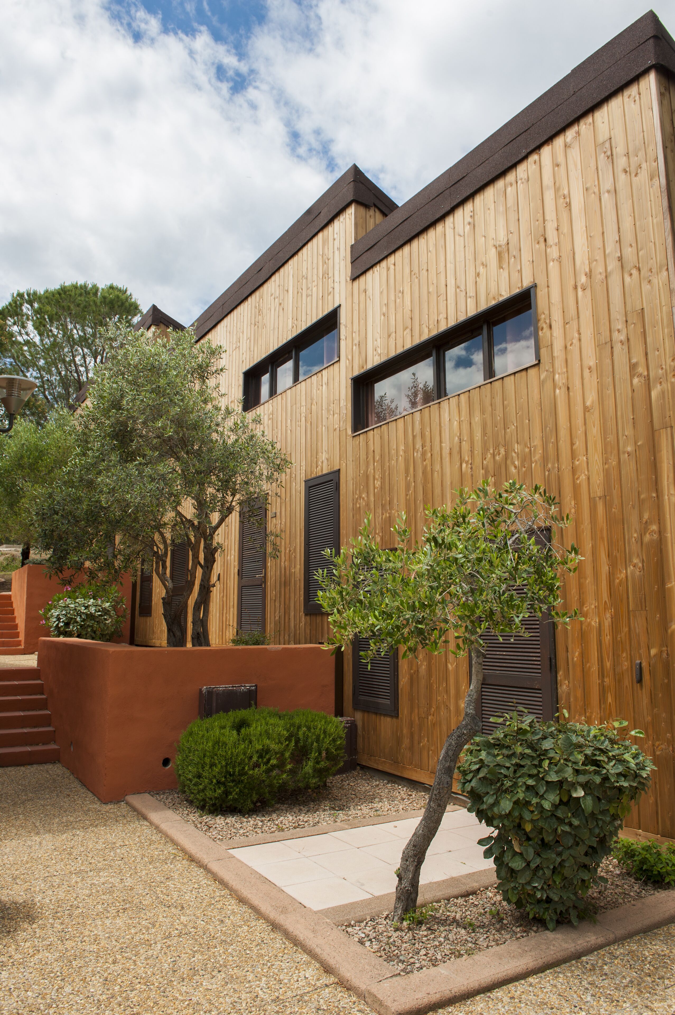 A modern building with wooden exterior walls, large windows, and a well-maintained garden with various shrubs and a tree.