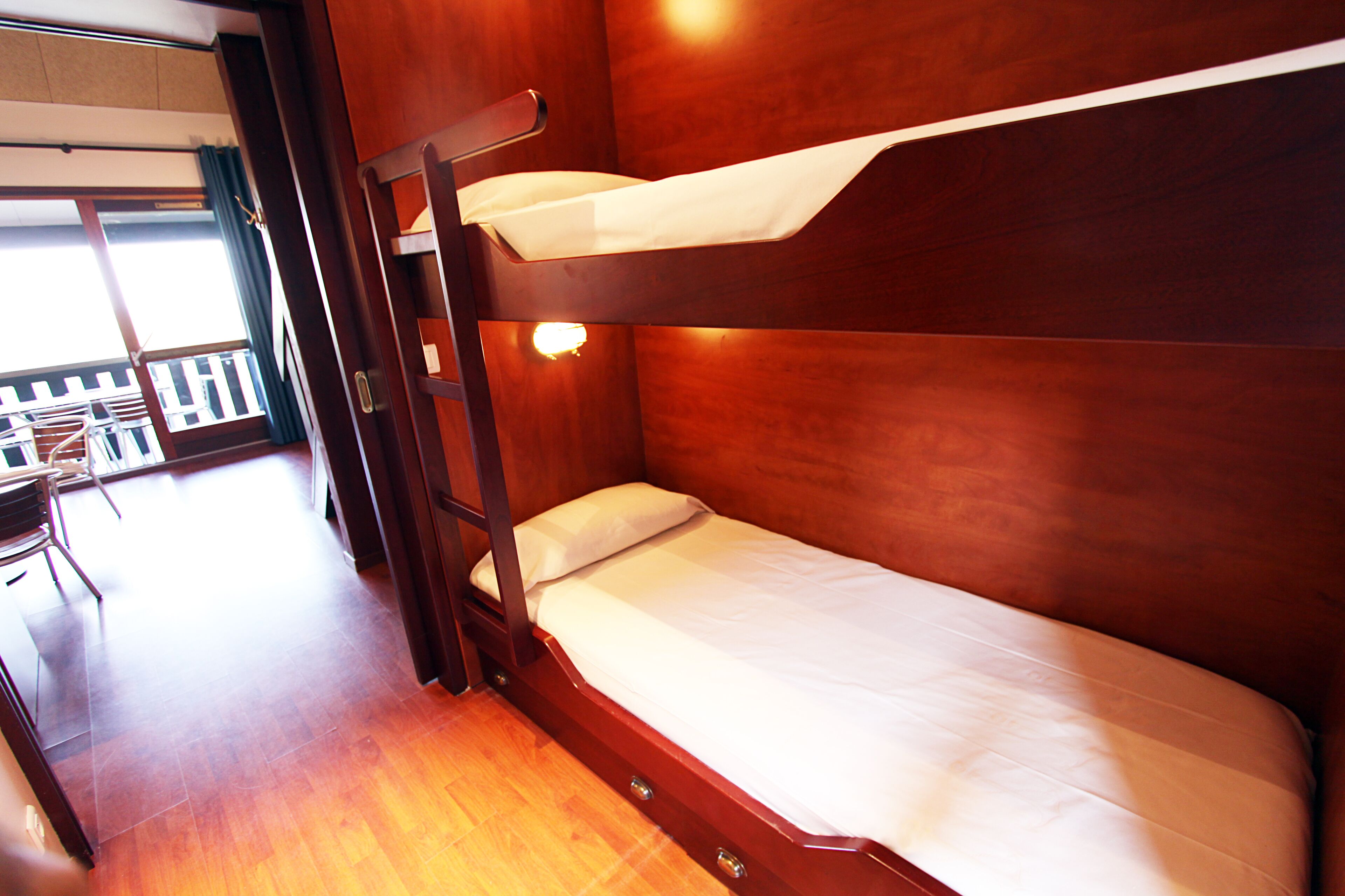 Een stapelbed met houten afwerking, een klein bureau met stoel, en uitzicht op een balkon met een hekwerk.