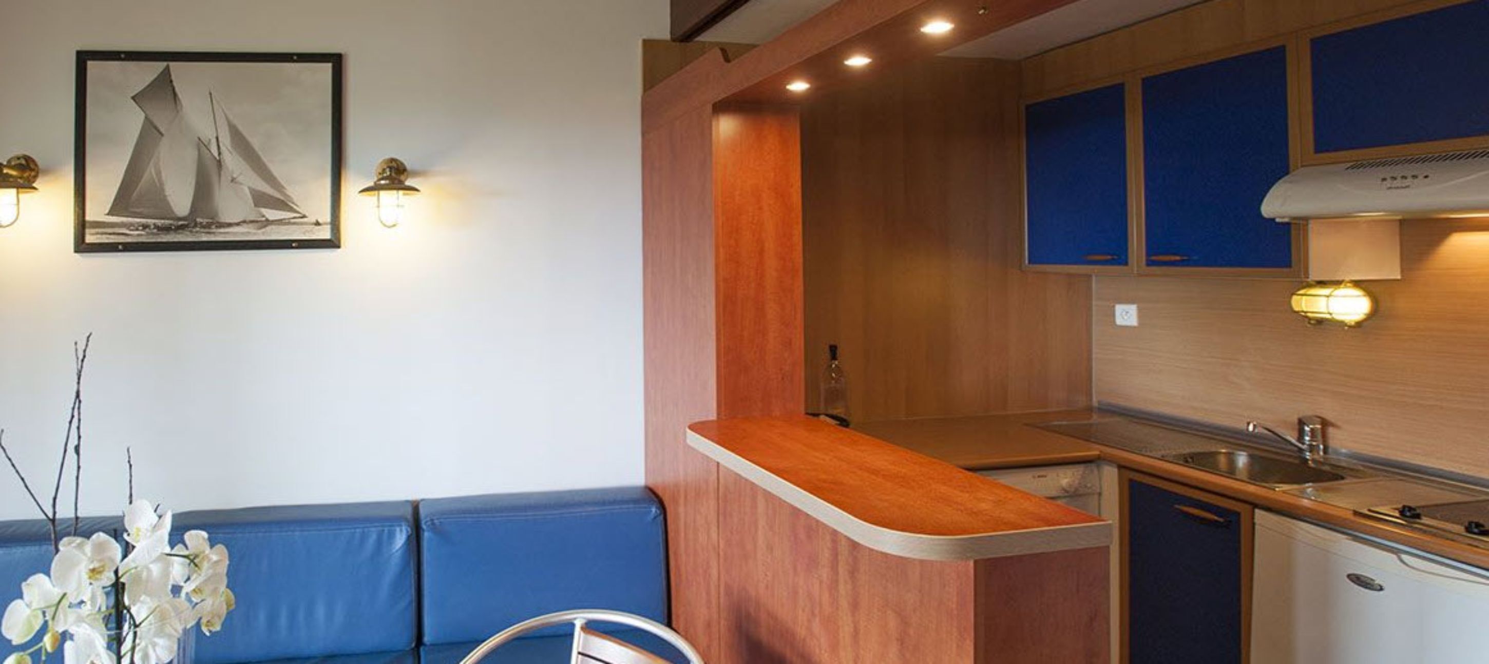Une cuisine compacte avec un plan de travail en bois, un évier et des placards. On y trouve un canapé bleu et une petite table avec une chaise. Un tableau encadré représentant des voiliers est accroché au mur.