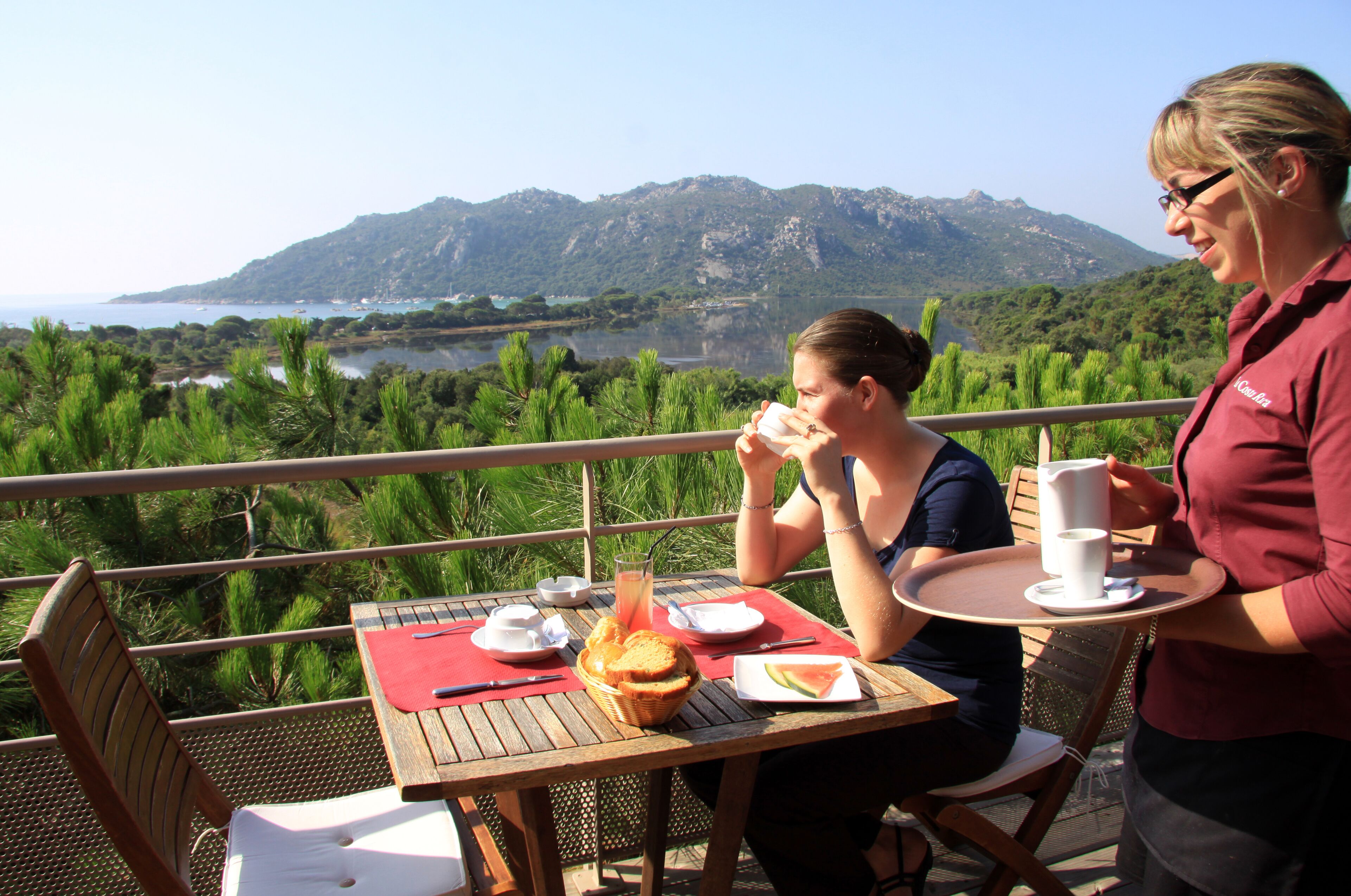 Una mesa de comedor con vista a un lago y montañas, una cesta de pan y un vaso de jugo.