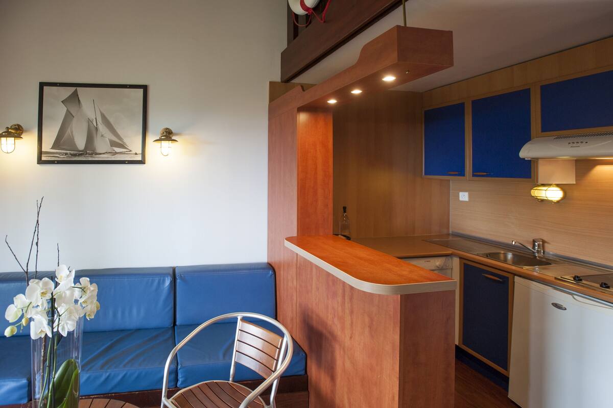A compact kitchen with a wooden countertop, a sink, and a built-in stove. There is a blue sofa and a chair in the living area, and a framed picture on the wall.