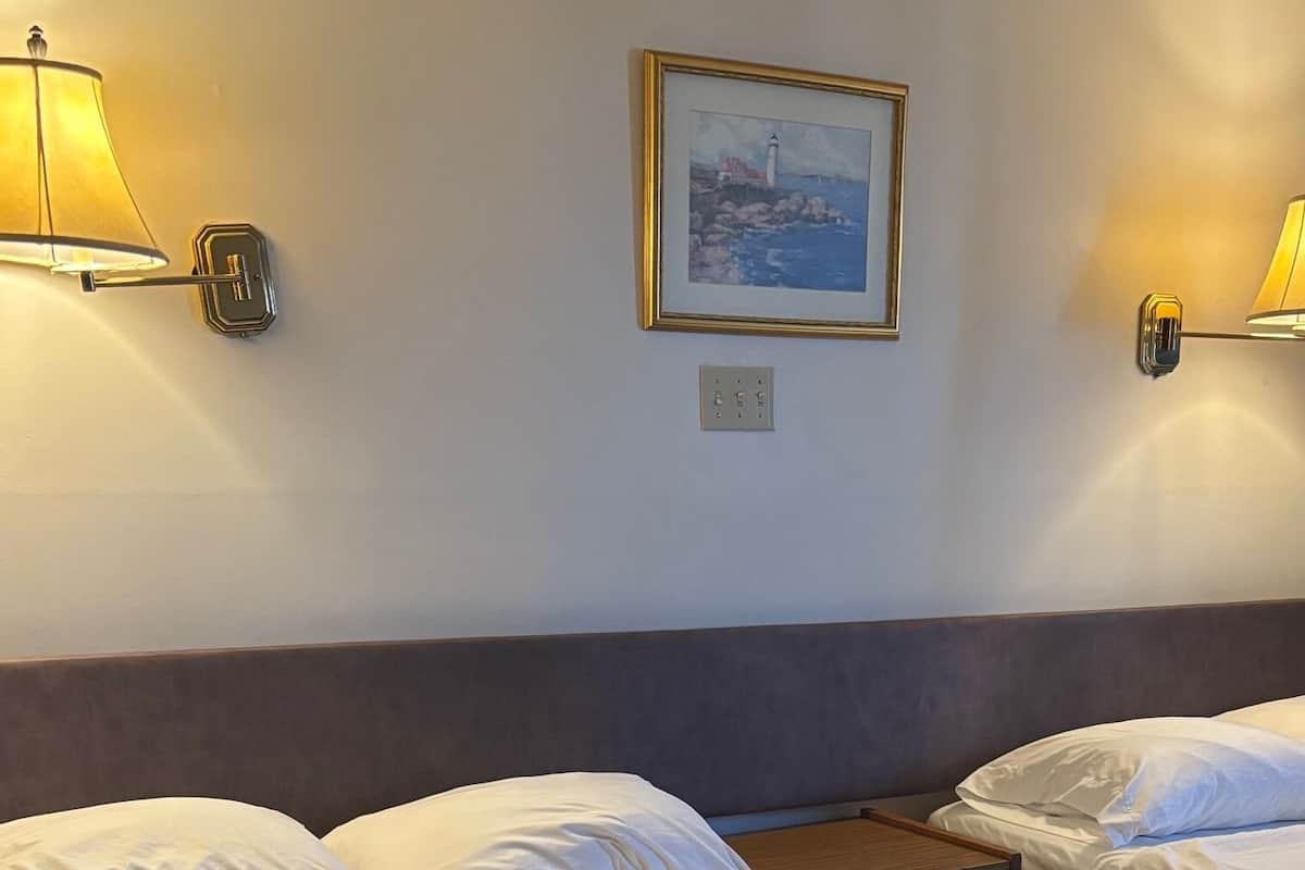 A hotel room with two beds, a painting on the wall, and two wall-mounted lamps.