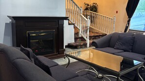 Un salon avec des canapés sombres, une table basse en verre, une cheminée et un escalier menant à un balcon.
