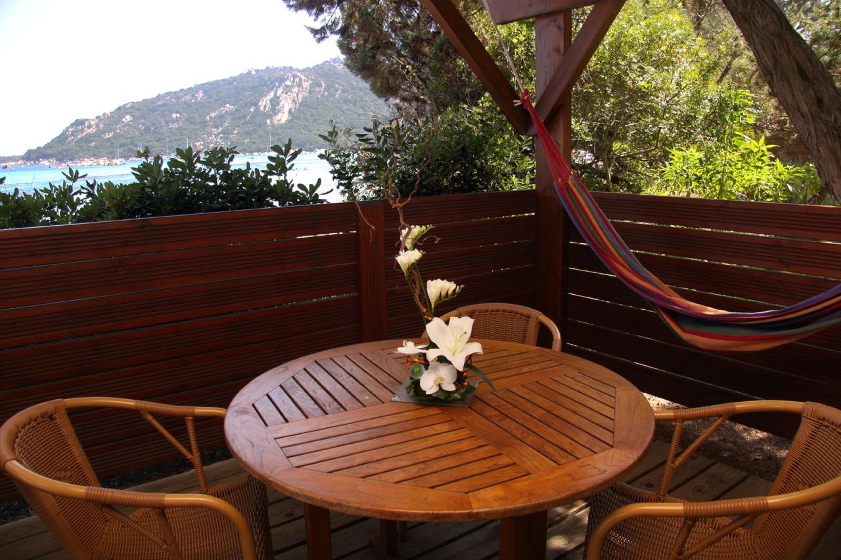 Una mesa de madera con sillas de mimbre y una hamaca, frente a un paisaje costero montañoso.