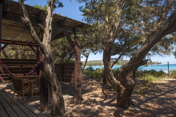 A wooden deck with a hammock, a table, and chairs, surrounded by trees and overlooking a body of water.