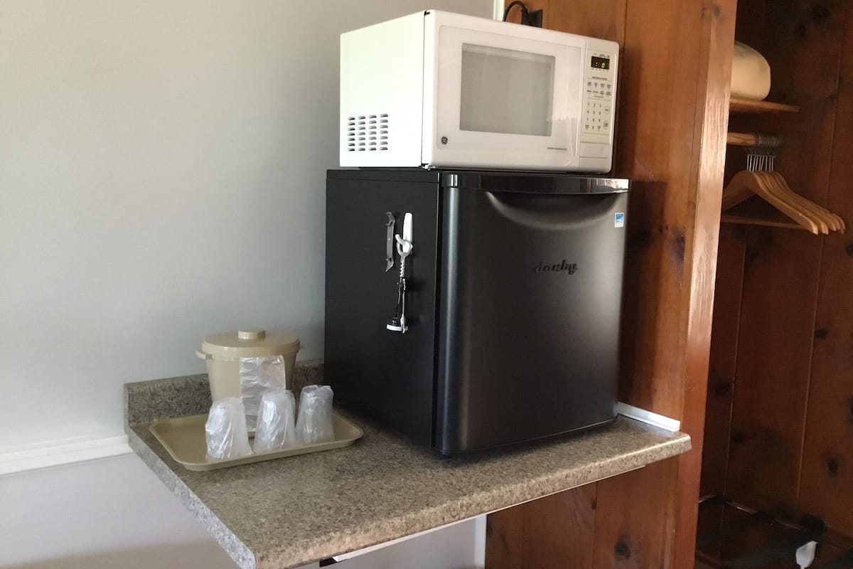 A compact refrigerator with a microwave on top, a small trash bin, and a tray with plastic bags on a granite countertop.