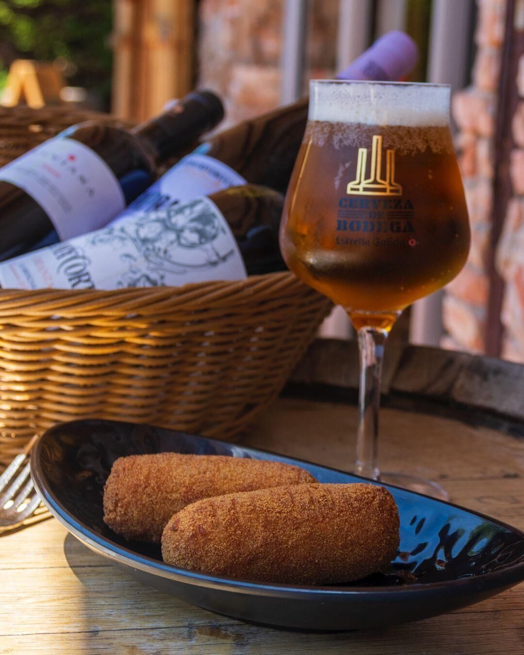 Un got de cervesa, una cistella de botelles de vi i dues croquetes sobre un plat.