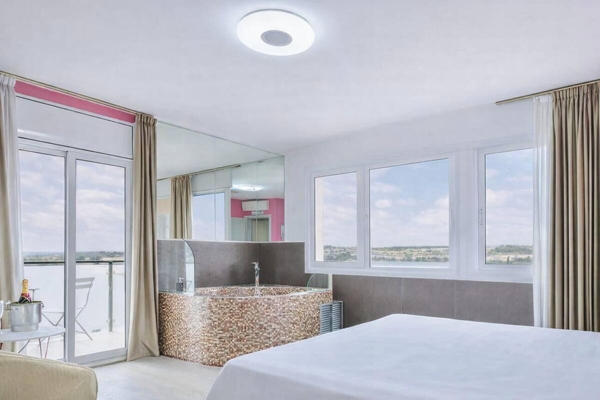 Habitación de hotel con una cama grande, un baño con bañera y un balcón con vistas.