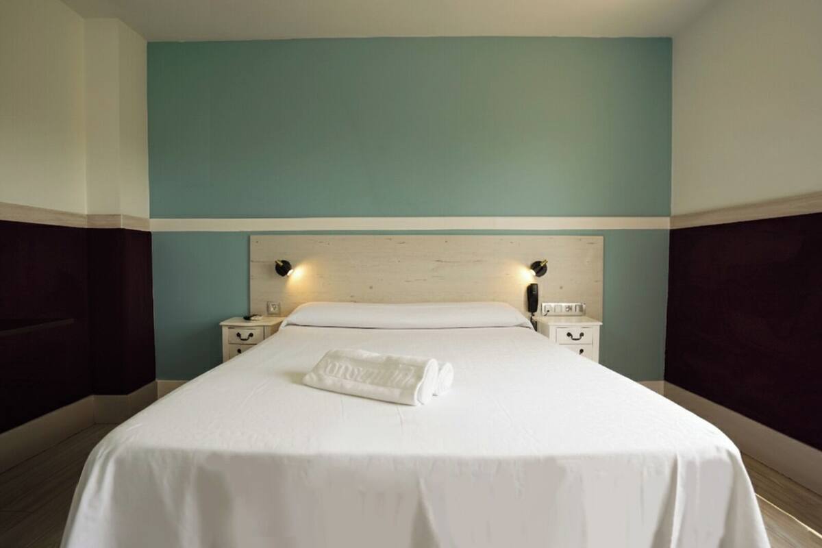 Una cama bien hecha con sábanas blancas y una toalla doblada, dos mesitas de noche con lámparas, y una pared color verde azulado.