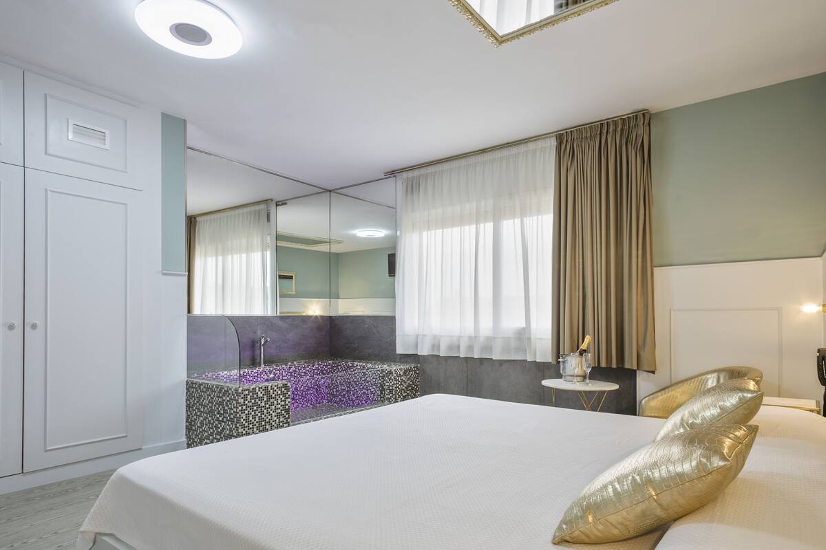 Una habitación de hotel con una cama grande, un baño con espejo y una mesita con una botella y vasos.